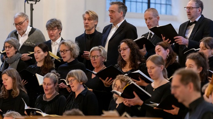 Ein Chor aus Männern und Frauen in schwarzer Kleidung, die von Notenblättern singen.