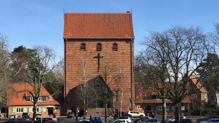 Ein rotes Backsteingebäude mit einem Kirchturm und einem Kreuz, umgeben von Bäumen und Autos.
