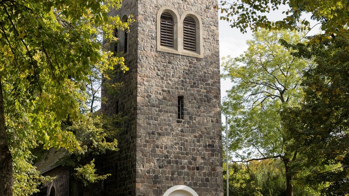 Der Kirchturm aus Stein mit Uhr und Fenster steht in einem Park.