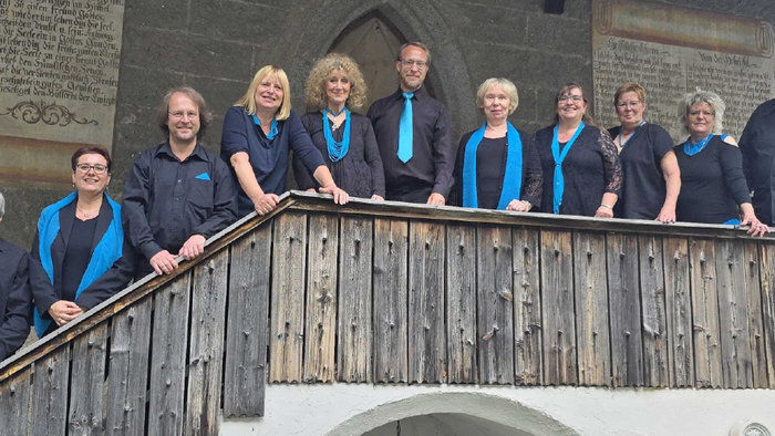 Der Spandauer Halbkreis: Zehn Sängerinnen und Sänger stehen auf einer hölzernen Empore vor der historischen Steinwand der Dorfkirche Staaken. Alle tragen schwarze Kleidung mit blauen Akzenten wie Schals, Krawatten oder Einstecktüchern und lächeln in die K