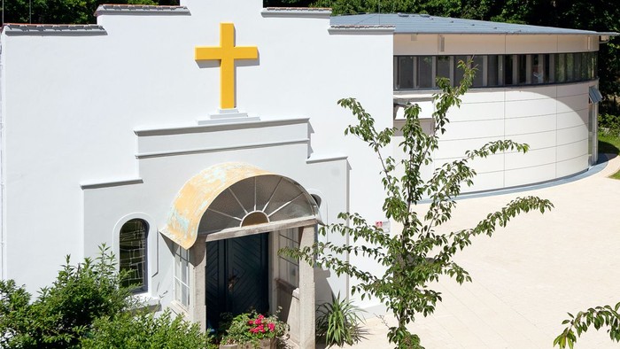 Das Bild zeigt eine Kirche mit einem gelben Kreuz und einem kleinen Garten.