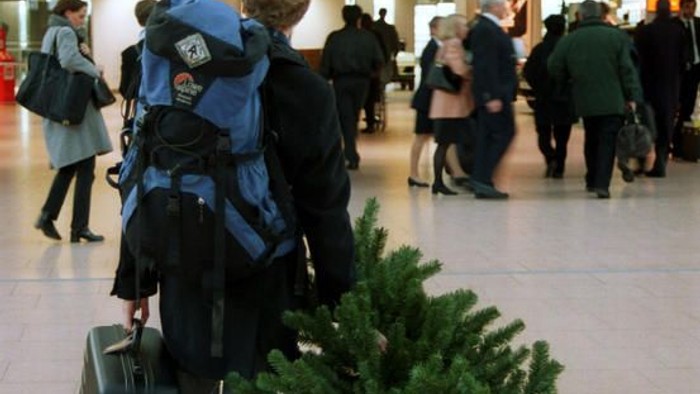 Reisender mit Gepäck und Rucksack steht in einem belebten Flughafen-Terminal in der Nähe eines kleinen künstlichen Weihnachtsbaums.