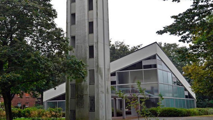 Das Bild zeigt eine moderne Kirche mit hohem Turm und Glasfront, umgeben von Bäumen.