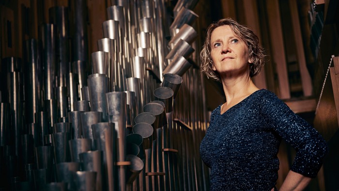 En kvinde står ved siden af et stort, indviklet rørformet musikinstrument, sandsynligvis et klokkespil eller et lignende slagtøjsopstilling.