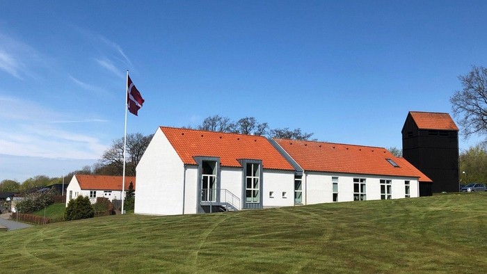 Hvidt hus med rødt tag og flagstang på grønne eng.