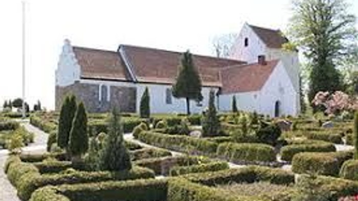 Et hvidt landhus med rødt tag og en formet have med grønne planter og træer.