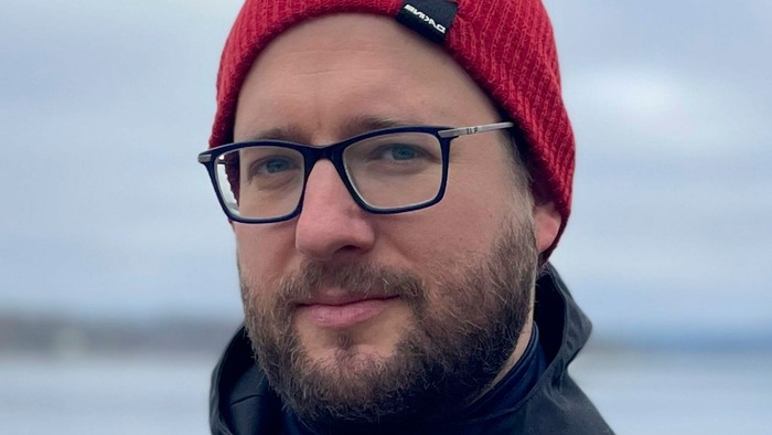 Mann mit Brille und roter Mütze, der draußen lächelt.