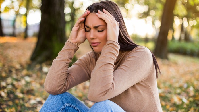 Eine junge Frau sitzt im Park und hält ihren Kopf, scheinbar in Gedanken versunken oder besorgt.