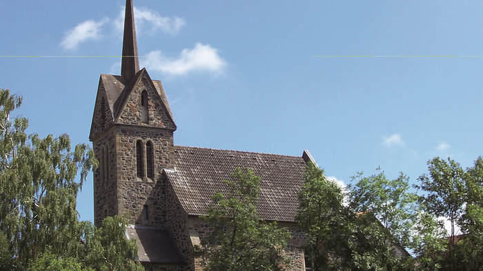 Kirchengebäude mit hohem Turm und spitzem Dach, umgeben von Bäumen unter blauem Himmel