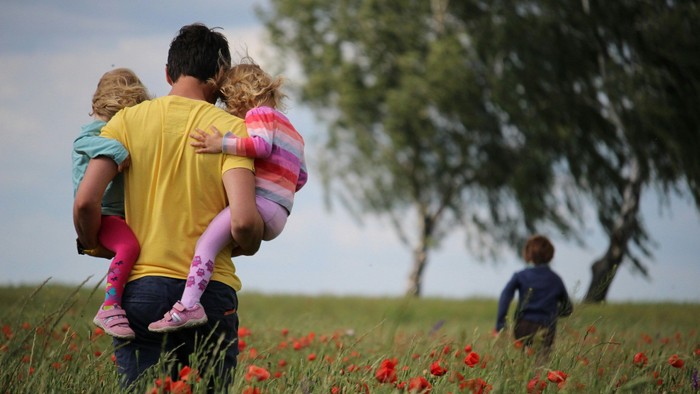 Ein Mann trägt zwei Kinder auf seinen Schultern, während ein drittes Kind im Hintergrund in einem Feld mit roten Blumen spielt.