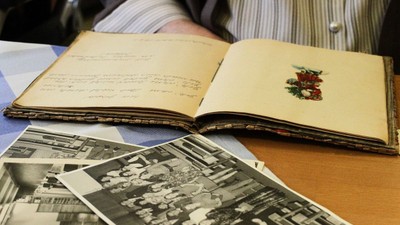 Ein aufgeschlagenes Buch mit handgeschriebenem Text und einer Stickerei, daneben liegen alte Schwarz-Weiß-Fotos