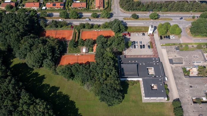 Luftfoto af tennisbaner omgivet af grønne områder, bygninger og en parkeringsplads nær et boligkvarter.