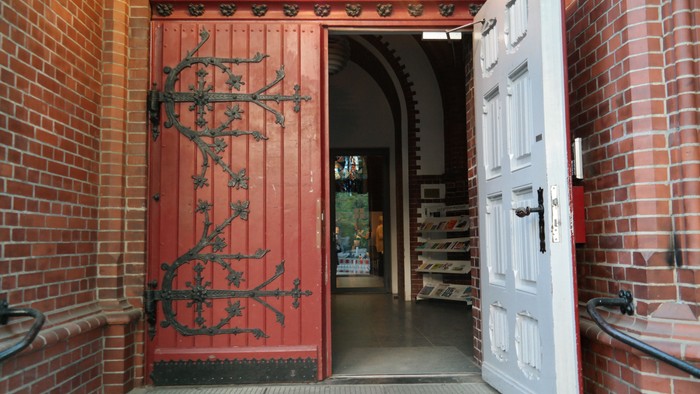 Ein rotes Tor mit schwarzem Eisenwerk ist in einem Ziegelgebäude eingebaut, daneben eine weiße Tür.