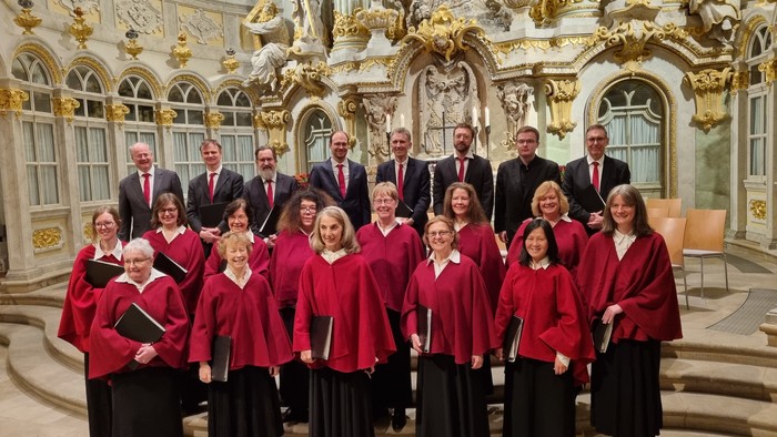 Ein Chor in roten Roben und Männer in Anzügen posieren gemeinsam im Inneren einer prächtigen, goldverzierten Kirche.