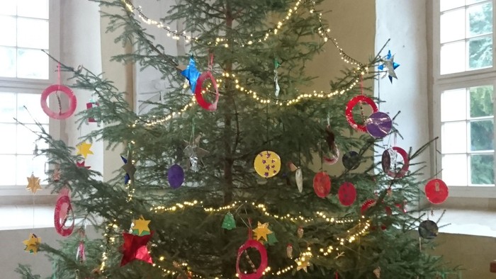 Ein Weihnachtsbaum mit bunten Kugeln und Lichtern steht vor einem Fenster.