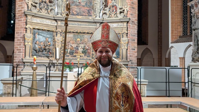 Geistlicher in religiöser Kleidung mit Stab vor einem prunkvollen Altar.