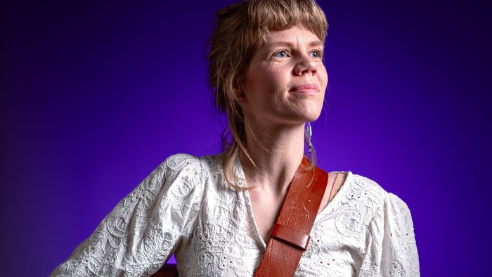 En person holder en guitarrem, mens de har en bluse med blonde og bukser med mønster på foran en lilla baggrund.