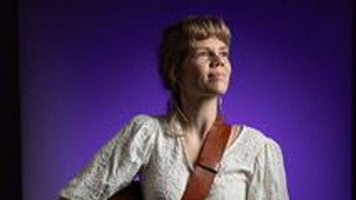 En person, der holder en guitar, iført hvid skjorte og mønstrede bukser, mod en lilla baggrund.