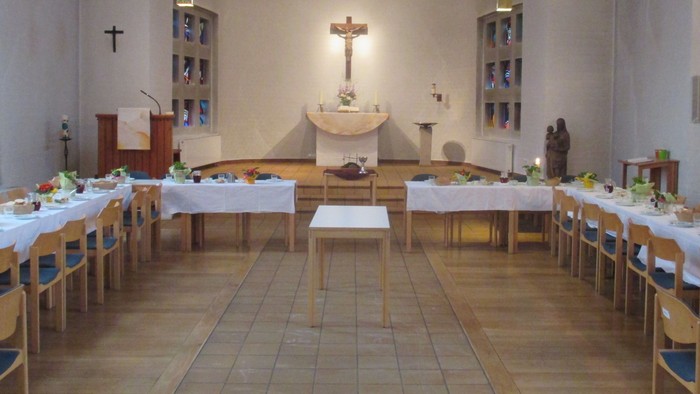 Ein großer Raum mit mehreren Tischen und Stühlen, die mit weißen Tischdecken und Blumen gedeckt sind, und einem Altar mit Kreuz in der Mitte