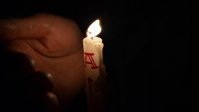 Eine Hand hält eine angezündete Kerze im Dunkeln.