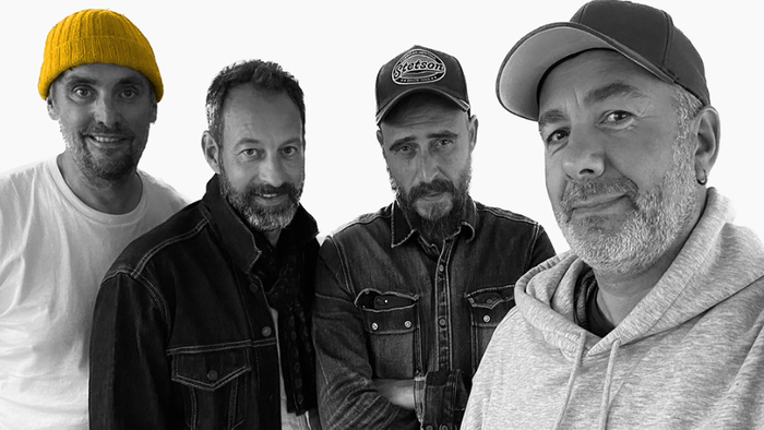 Fire mænd, der poser sammen, tre i farver og en i sort-hvid.