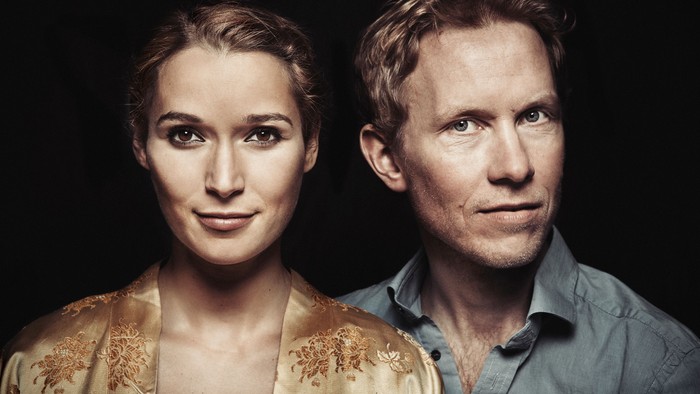 To personer står tæt sammen mod en mørk baggrund og udstråler en alvorlig og afmålt holdning.