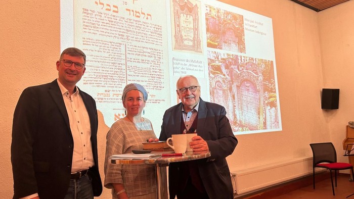 Drei Personen stehen in der Nähe einer projizierten Anzeige, eine hält einen Becher.