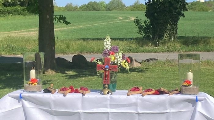 Gedenktisch im Freien mit Blumen, Kerzen und Speisen unter einem Baum in einer ruhigen Landschaft.