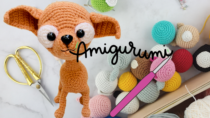 Ein gehäkelter Hund, schere mit goldenem Griff, Bunte Wolle eine Häkelnadel und Ande ein Schriftzug Amigurumi der aus dem Maul vom Hund kommt.
