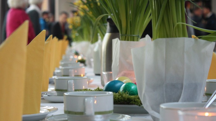 Ein festlich gedeckter Tisch mit weißen Tellern, Tassen und grünen Ostereiern, begleitet von gelben Servietten und Topfpflanzen