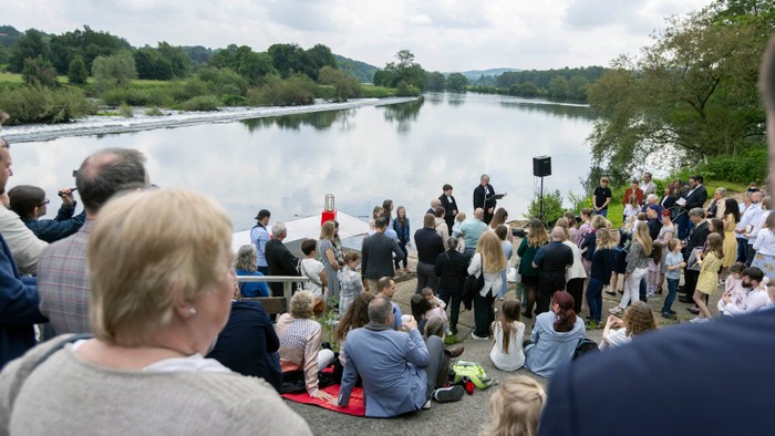 Eine Gruppe von Menschen versammelt sich am Ufer eines Flusses, einige sitzen, andere stehen und unterhalten sich.