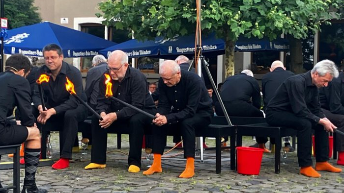 Gruppe von Männern in schwarzer Kleidung mit gelben Schärpen, die im Freien eine synchronisierte Balance-Übung auf Stangen vorführen.