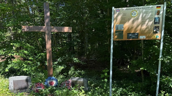 Ein rustikales Holzkreuz in einem bewaldeten Gebiet mit Informationstafeln und nahen Grabsteinen.
