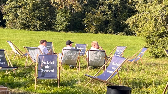 Drei Personen sitzen auf Klappstühlen im Gras, jeder hält ein Schild mit der Aufschrift 