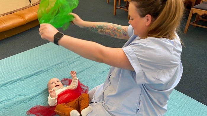 En kvinde med tatovering på arm og klokke på hånd, holder en grøn plastflaske over et babyudstyr.