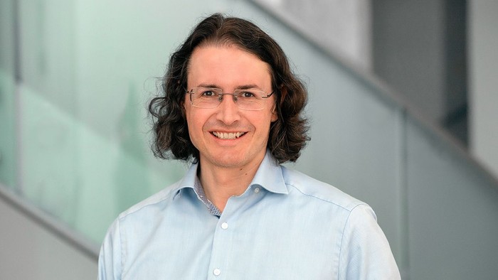 Ein lächelnder Mann mit Brille und langen Haaren, der ein hellblaues Hemd mit Knöpfen trägt, drinnen.