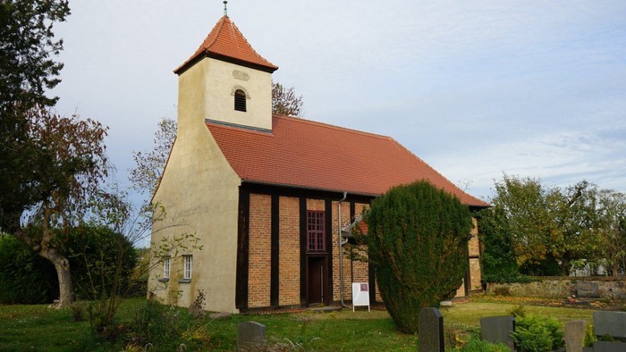 Kirche mit Ziegeldach, rechteckigem Turm und Fachwerkfassade, umgeben von Bäumen und Grabsteinen.