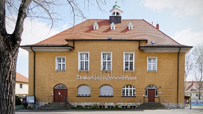 Frontalansicht vom Gemeindehaus Kreuz. Zweigeschossiger Bau mit ockerfarbener Fassade, Walmdach und kleinem mittigen Türmchen mit Glocke.