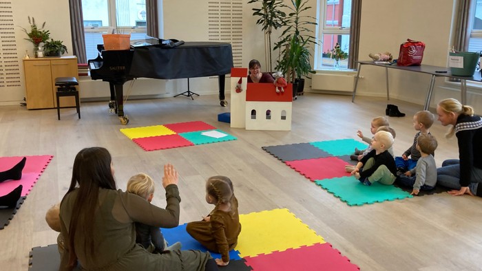 En gruppe børn og voksne sidder på farverige mattebrædder i et rum med piano og dække.