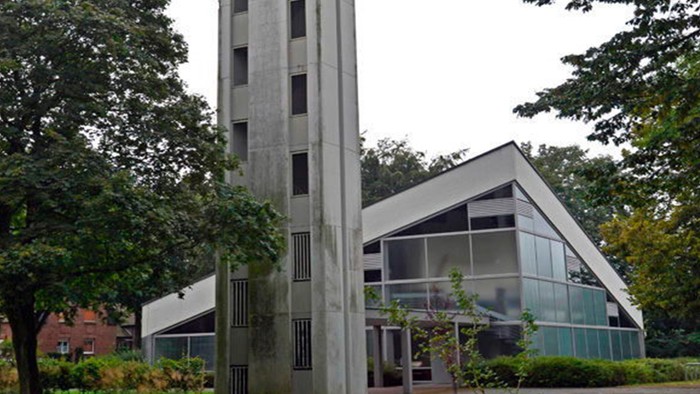 Das Bild zeigt eine moderne Kirche mit hohem Turm und Glasfront, umgeben von Bäumen.