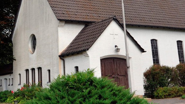 Weiße Kirche mit hohem Turm und braunem Dach, umgeben von Bäumen und Büschen