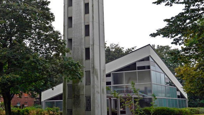 Das Bild zeigt eine moderne Kirche mit hohem Turm und Glasfront, umgeben von Bäumen.