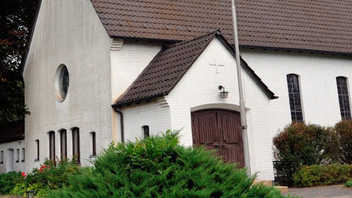 Weiße Kirche mit hohem Turm und braunem Dach, umgeben von Bäumen und Büschen