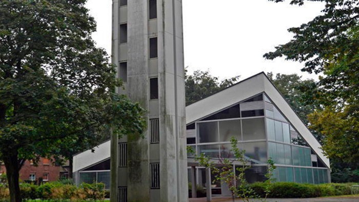 Das Bild zeigt eine moderne Kirche mit hohem Turm und Glasfront, umgeben von Bäumen.