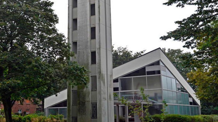 Das Bild zeigt eine moderne Kirche mit hohem Turm und Glasfront, umgeben von Bäumen.