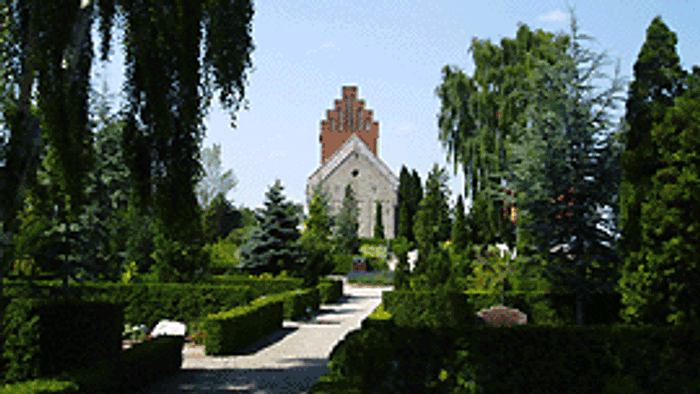 En lille kirke med røde murværker og hvidt tag, omgivet af træer og buske.