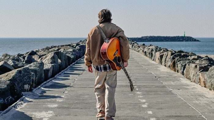 En person går langs en havnebro med en akustisk guitar og kigger ud over havet.