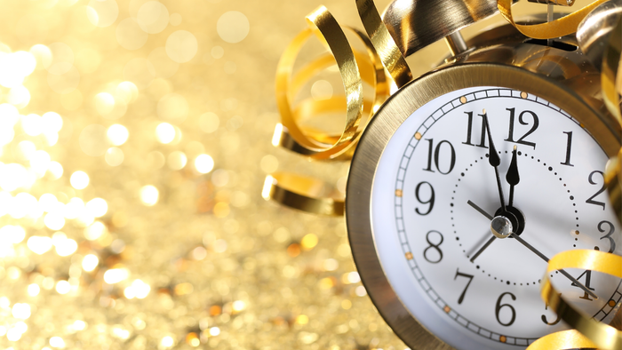 Ein goldener Wecker mit schwarzen Zeigern steht auf einer glitzernden goldenen Oberfläche, umgeben von goldenen Konfettibändern