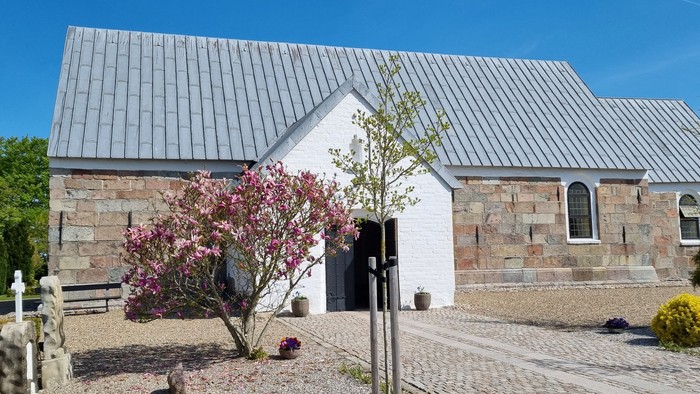 Et moderne hus med metaltag og stenfacade, omgivet af træer og blomster.