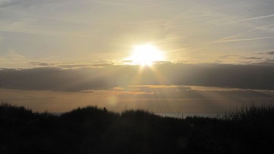 „Die untergehende Sonne scheint hell durch verstreute Wolken über eine silhouettierte Landschaft.“